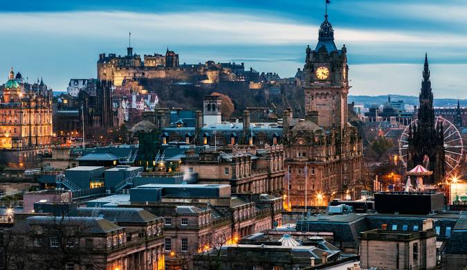 🏰 **Edimburgo, Escócia** – Uma cidade que combina história, cultura e beleza arquitetônica. Na imagem, vemos o icônico **Castelo de Edimburgo** no topo da colina, iluminado ao entardecer, com a **Balmoral Clock Tower** em destaque e a roda-gigante do **Princes Street Gardens** ao fundo. Uma paisagem que reflete a atmosfera mágica da capital escocesa.